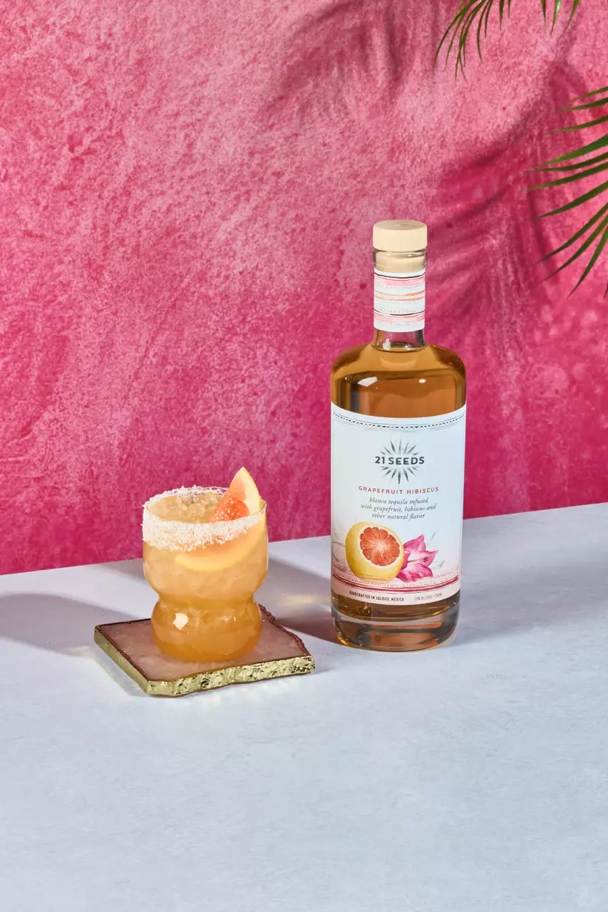 A bottle of 21 Seeds Grapefruit Hibiscus tequila sits next to a cocktail garnished with a grapefruit wedge on a pink and white surface.