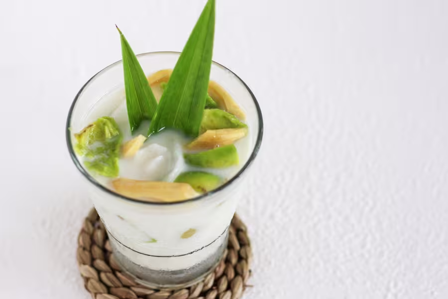 A glass of coconut milk dessert with jackfruit, avocado, ice, and pandan leaves, placed on a woven coaster against a white background.