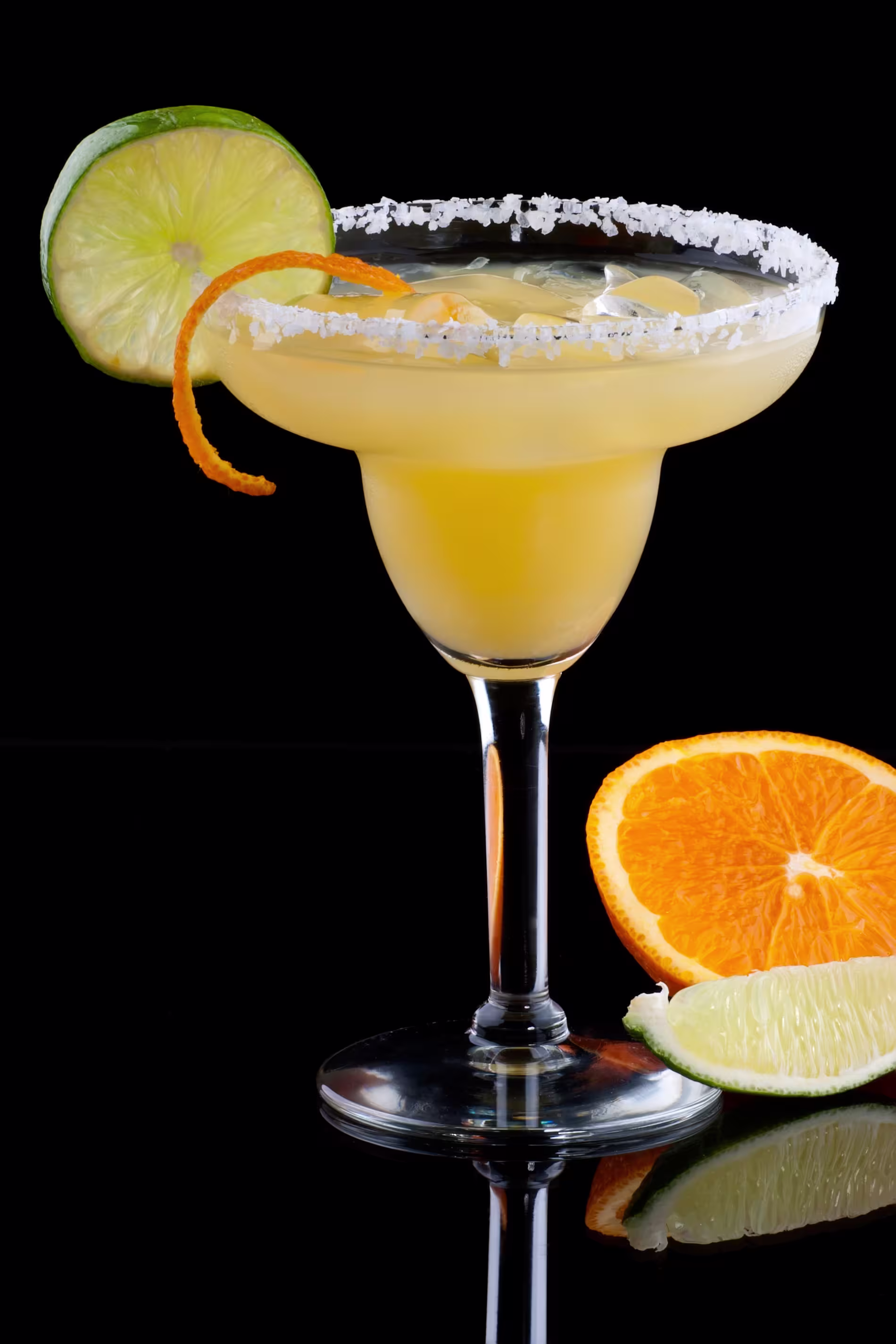 A margarita in a salt-rimmed glass garnished with a lime slice and orange twist, with orange and lime wedges beside the glass, against a black background.