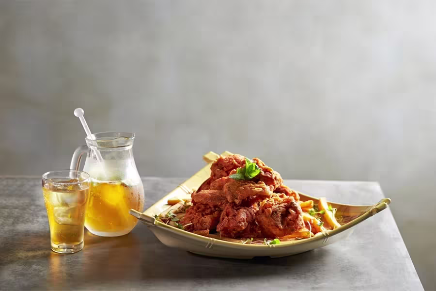 A plate of fried chicken garnished with herbs is placed beside a glass pitcher and a glass of iced tea on a gray table.