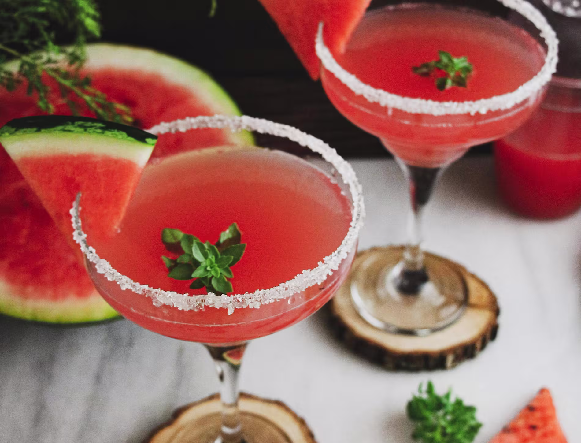 Two cocktail glasses with pink drinks, garnished with watermelon slices, sugared rims, and mint sprigs, set on wooden coasters with a watermelon slice in the background.