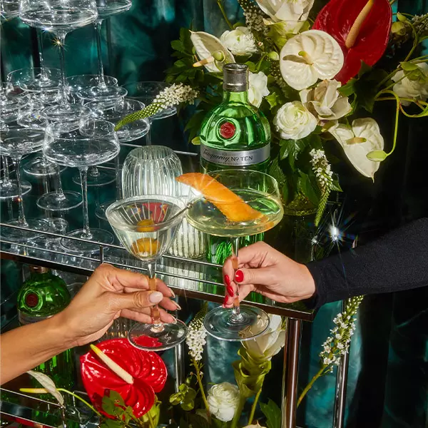 Two hands clink cocktail glasses in front of a bar cart with green bottles, stacked glassware, and an arrangement of red and white flowers.