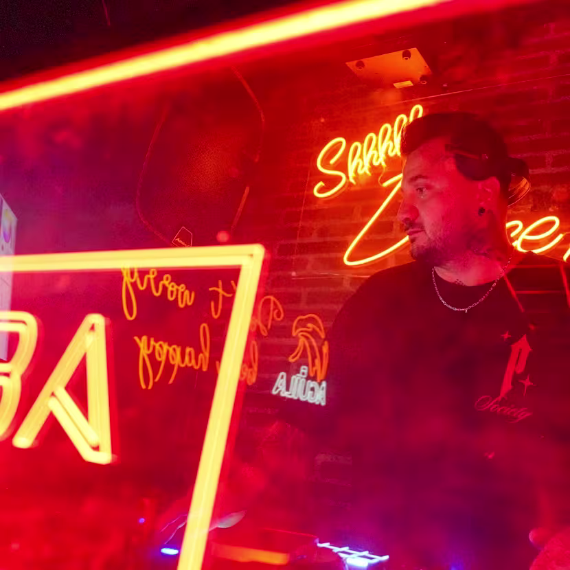 Malaba: Ambiente vibrante en tonos rojos neón, con un DJ al mando de la cabina, rodeado de luces llamativas y letreros que dan un toque urbano y moderno al espacio."