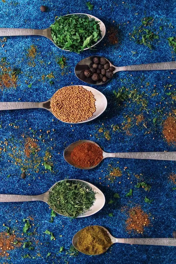 Six metal spoons filled with various spices and herbs are arranged in a vertical line on a blue textured surface, with scattered spice remnants around them.