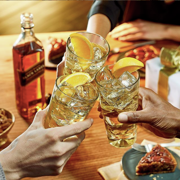 La imagen muestra un brindis con tres vasos con cócteles preparados con whisky Johnnie Walker y de fondo, una botella de whisky Johnnie Walker Black Label y distintos platos de comida.