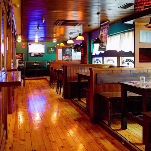 La imagen muestra el interior del Irish Corner con cabinas y mesas de madera. Hay letreros de marcas de whisky, y elementos decorativos con temática irlandesa.