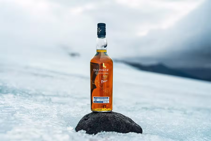 A bottle of Talisker whisky stands on a rock surrounded by ice, with a cloudy sky and distant mountains in the background.
