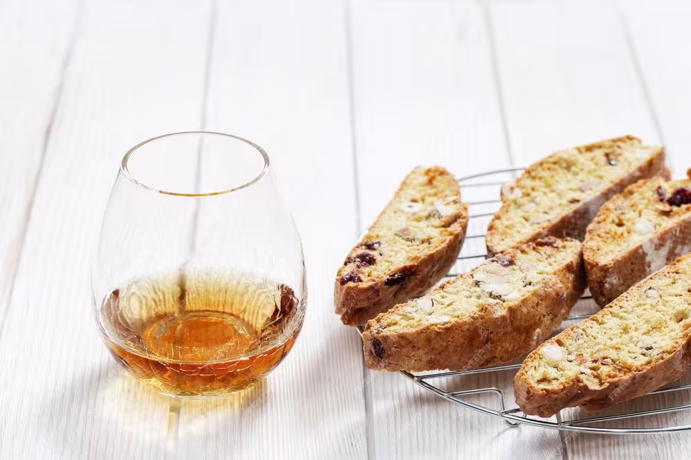 A glass of amber-colored liquor sits next to several pieces of biscotti on a cooling rack, all on a light wooden surface.