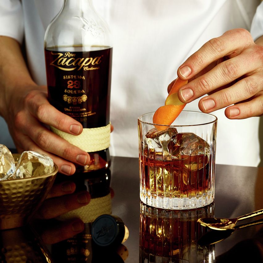 La imagen muestra a una persona agarrando una botella de ron Zacapa 23 y colocando una rodaja de naranja en un vaso que contiene el cóctel Old Fashioned.