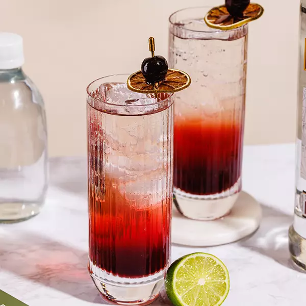 Two tall glasses filled with a red and clear layered drink over ice, garnished with a dried lime slice and cherry, with a halved lime and bottle nearby.