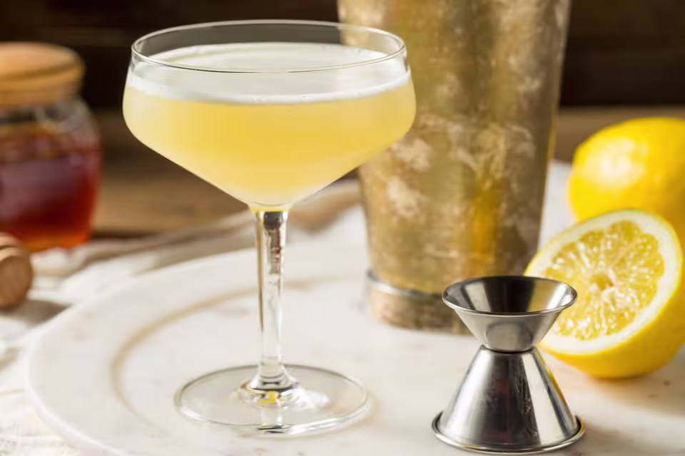 A yellow cocktail in a coupe glass sits on a tray with a metal jigger, a lemon half, a cocktail shaker, and a jar of honey in the background.