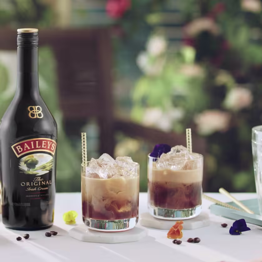 La imagen muestra una botella de licor de crema Baileys junto a dos vasos con cócteles preparados con este licor, una cafetera y otros elementos de coctelería.