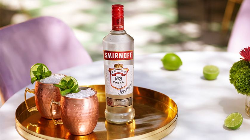 En la imagen se observan dos tazas de cobre con el cóctel Moscow Mule y una botella de vodka Smirnoff No. 21 sobre una bandeja dorada, con tres limones dispuestos alrededor.