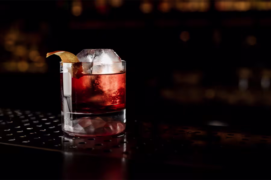 A glass of red cocktail with a large ice cube and an orange peel garnish sits on a perforated bar counter with a dark, blurred background.