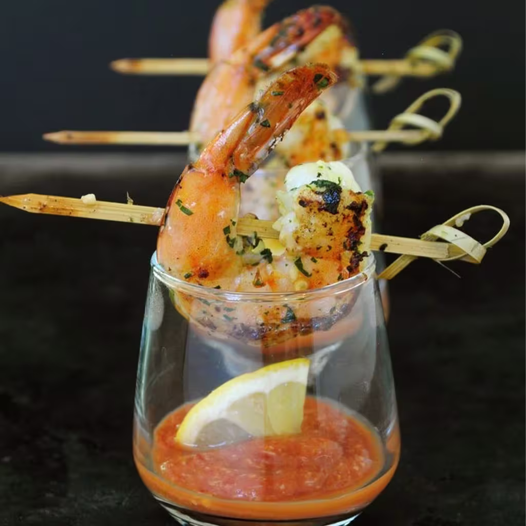 Three grilled shrimp skewers rest on the rims of glasses filled with cocktail sauce and lemon wedges, arranged in a row against a dark background.