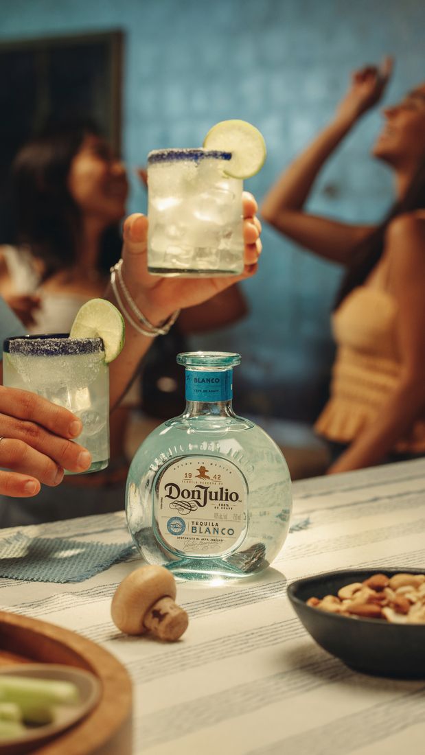 La imagen muestra una botella de tequila Don Julio Blanco, dos vasos rellenos del cóctel Margarita y, de fondo, dos personas.