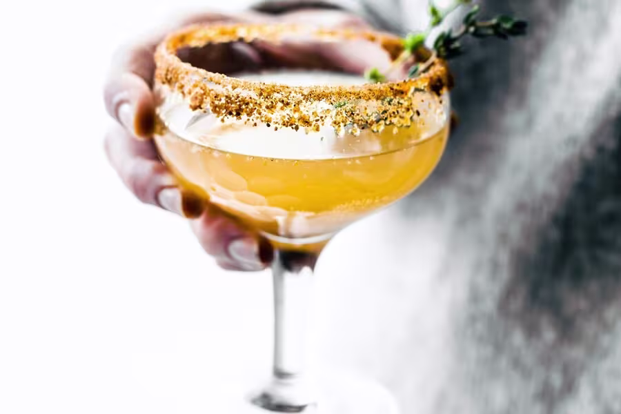 A hand holds a coupe glass filled with an orange cocktail, garnished with a spiced rim and a sprig of fresh herbs.