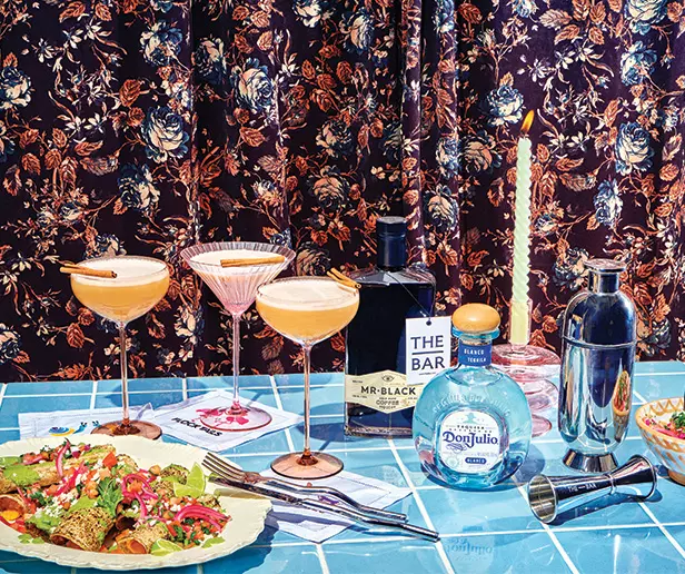 A tiled table with cocktails, a salad plate, cocktail shaker, bottles of Mr Black and Don Julio, a candle, bar tools, and floral curtains in the background.