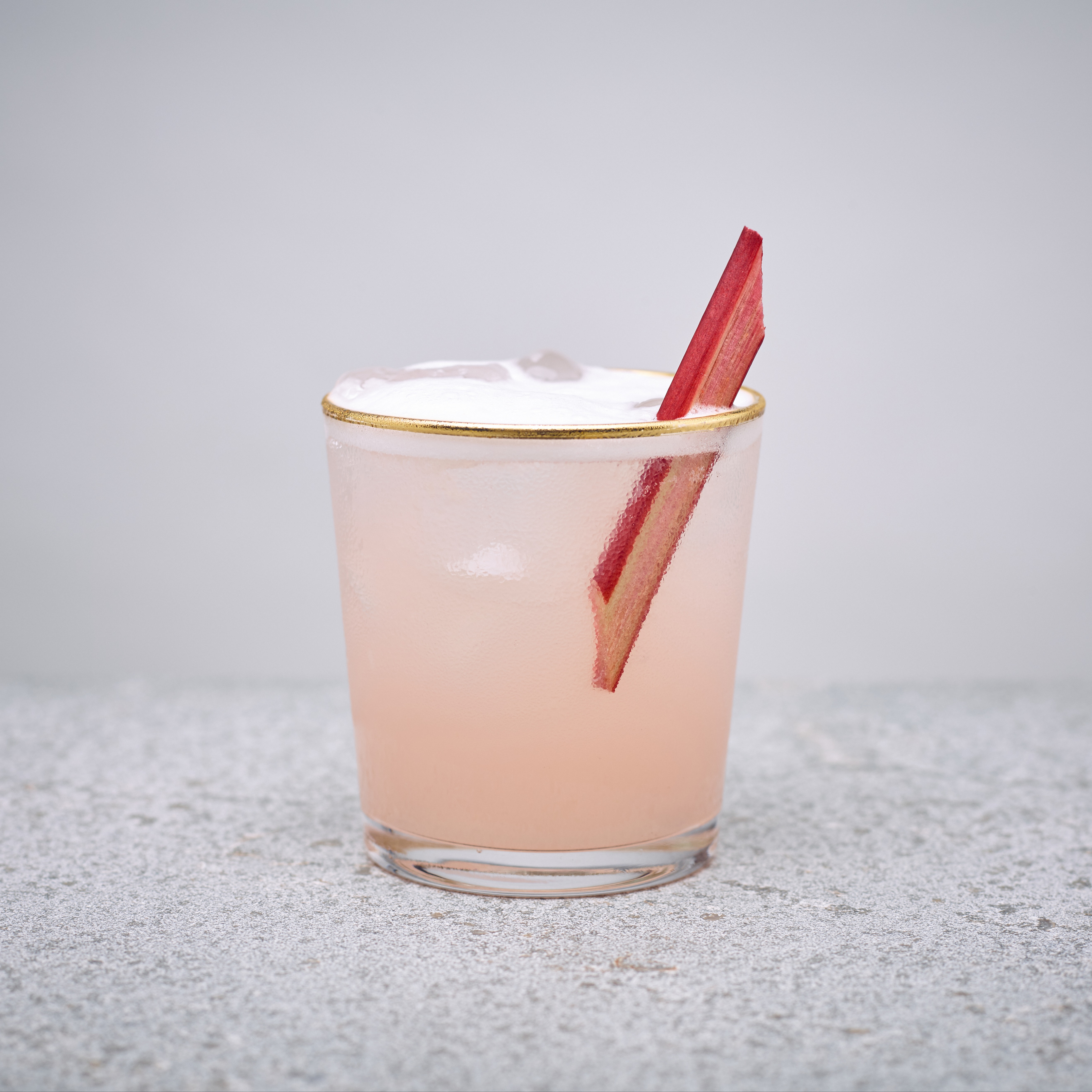 A rhubarb sour cocktail in a short glass with a sliced piece of rhubarb as garnish. 