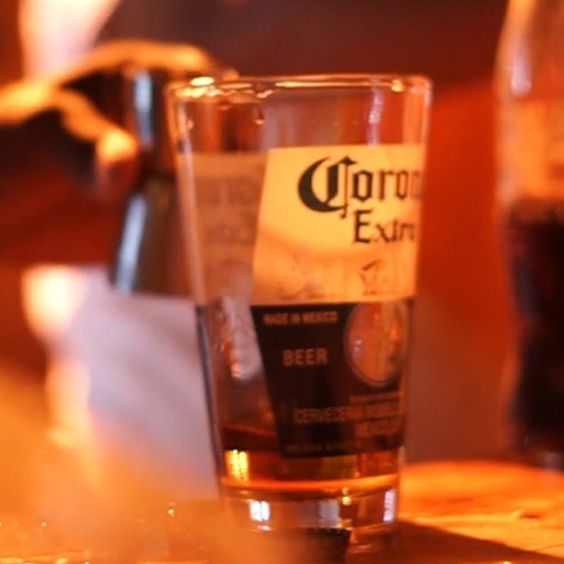 En la imagen se observa un vaso con las inscripciones de "Corona" sobre una mesa. En el fondo desenfocado se distinguen otros cócteles, un destapador, una bebida sin alcohol y el brazo de un hombre preparando un cóctel.