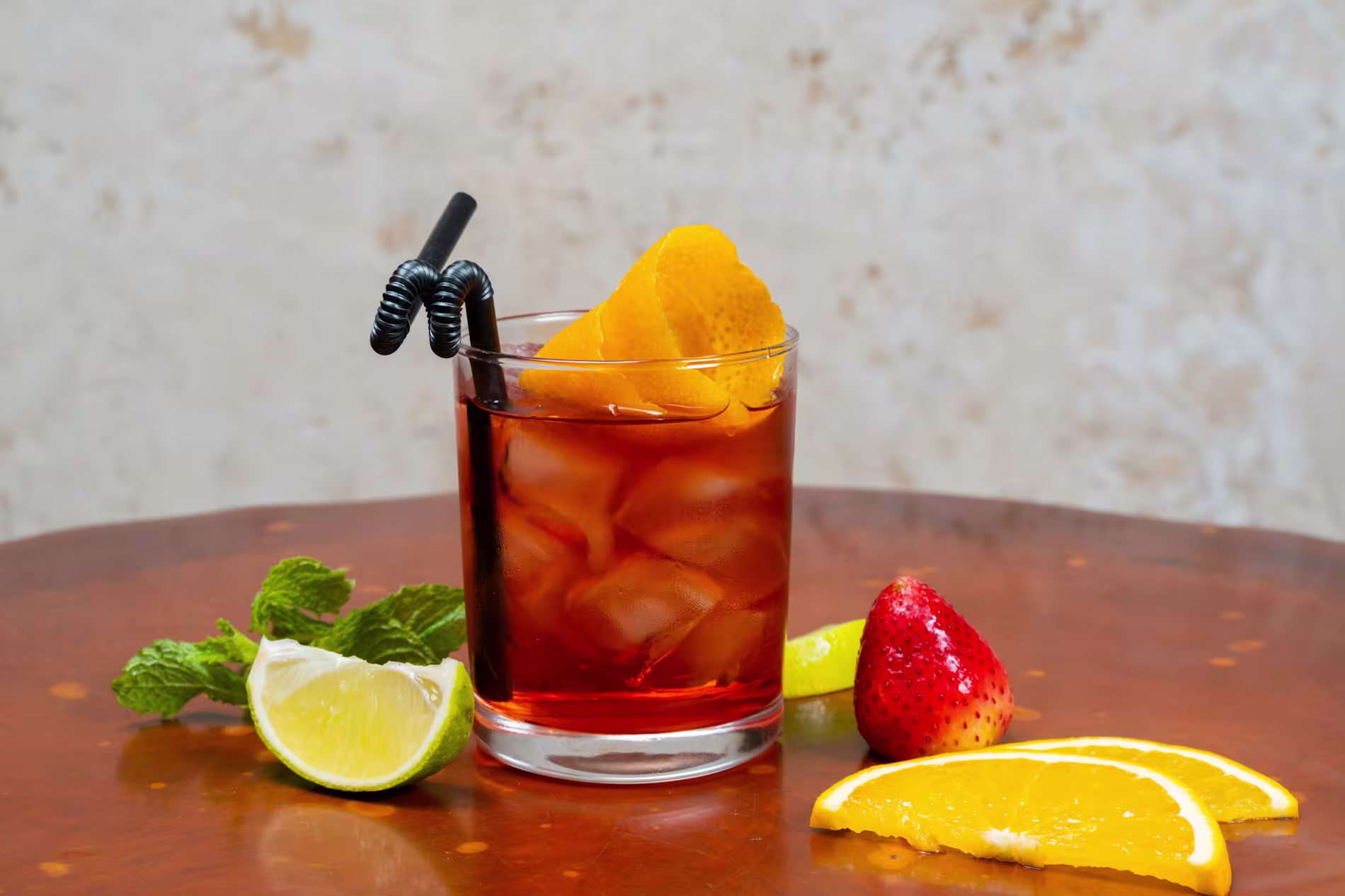 A glass of iced cocktail with an orange peel garnish, surrounded by lime, orange slices, a strawberry, and mint on a wooden surface.