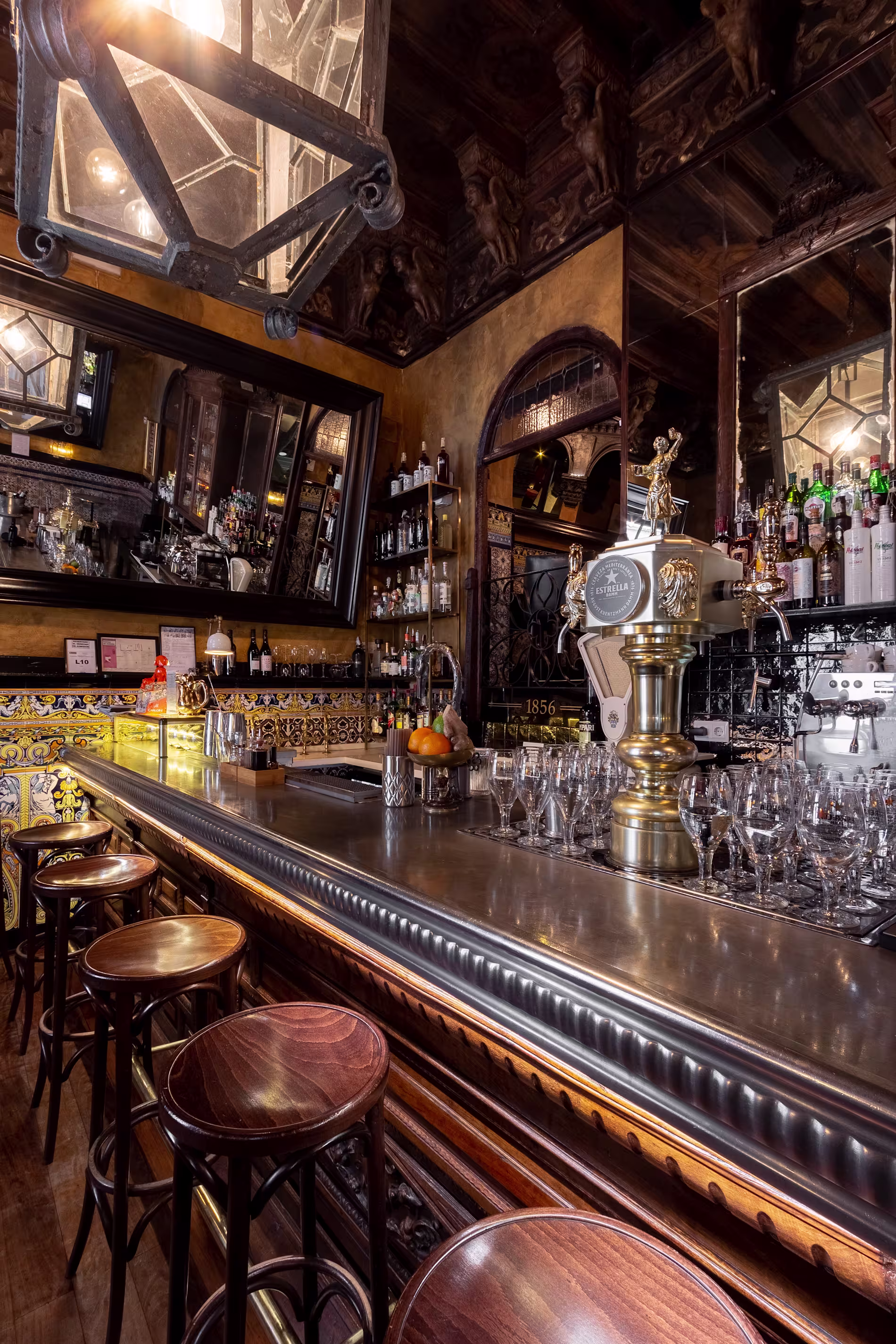 La imagen muestra el interior del Viva Madrid, condiseño clásico y ornamentado. Hay una barra de madera con taburetes altos frente a ella, y se puede ver una variedad de botellas y vasos. La pared del fondo tiene elementos decorativos y espejos.