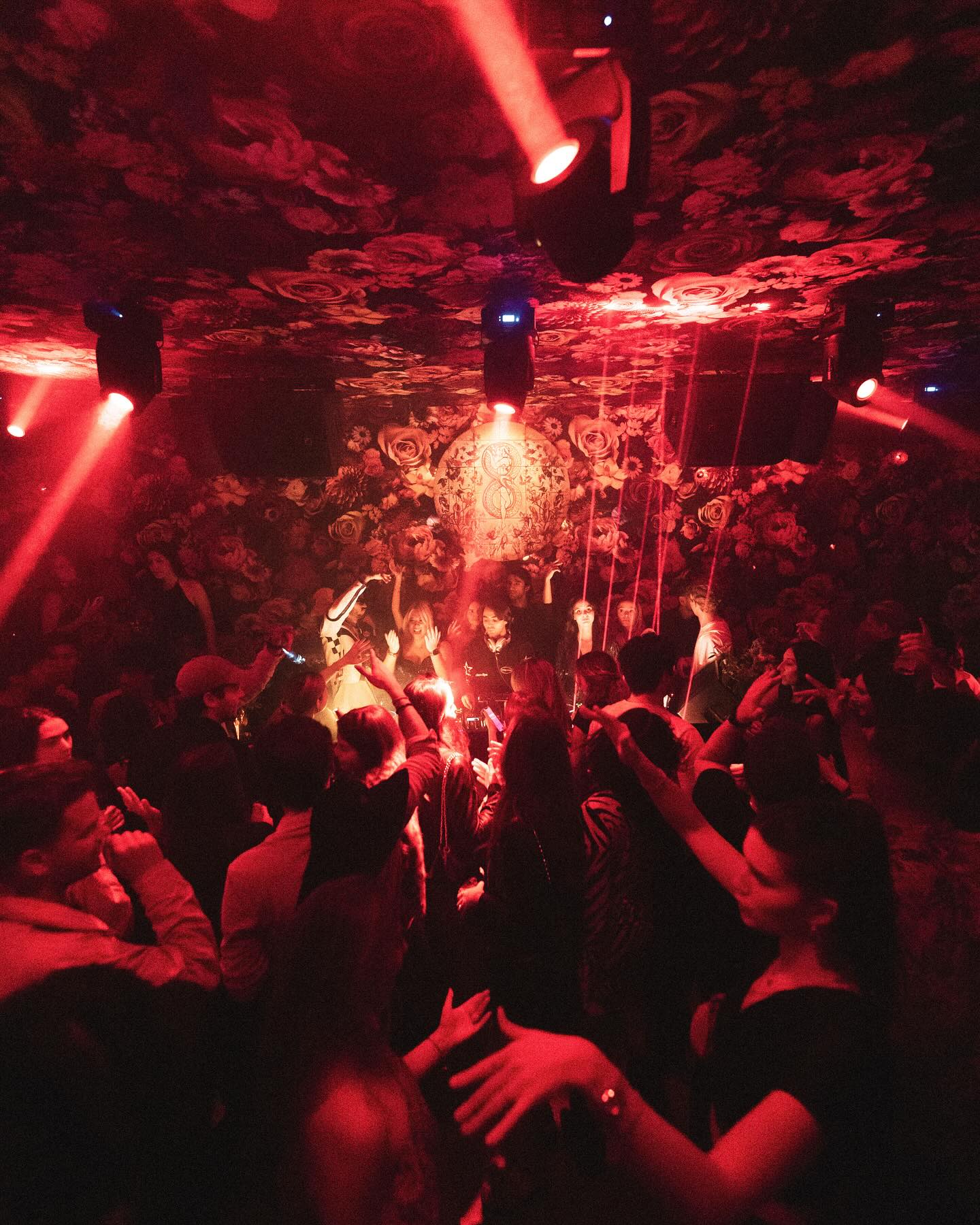 Personas bailando bajo una iluminación roja en el club Los Amantes, rodeados de un decorado floral que crea un ambiente único.