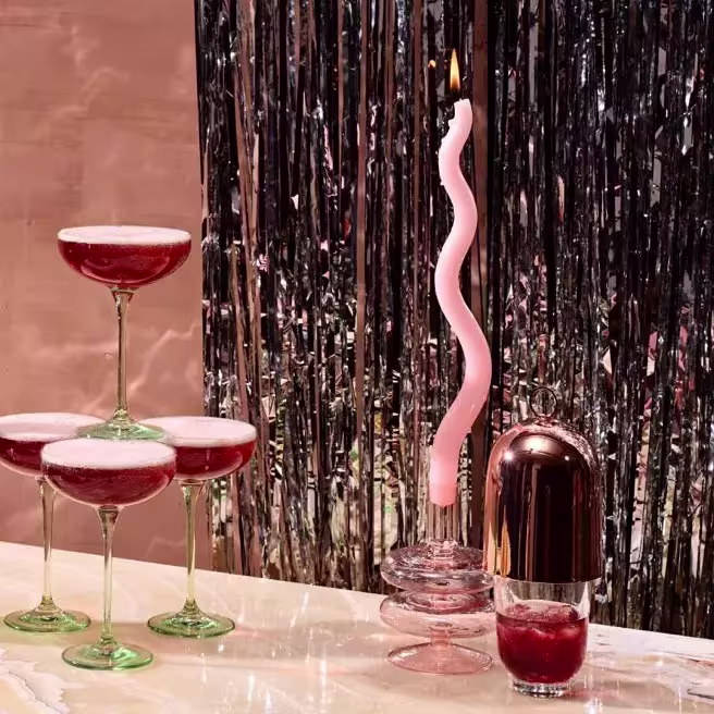 Four coupe glasses with a red drink are stacked in a pyramid beside a lit pink twisted candle and a drink in a glass with a metallic shaker, all on a table with a tinsel backdrop.