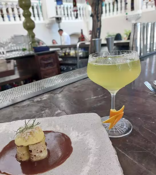 En la imagen se ve la barra del lugar en el exterior. Un cóctel y un pequeño plato apoyados.