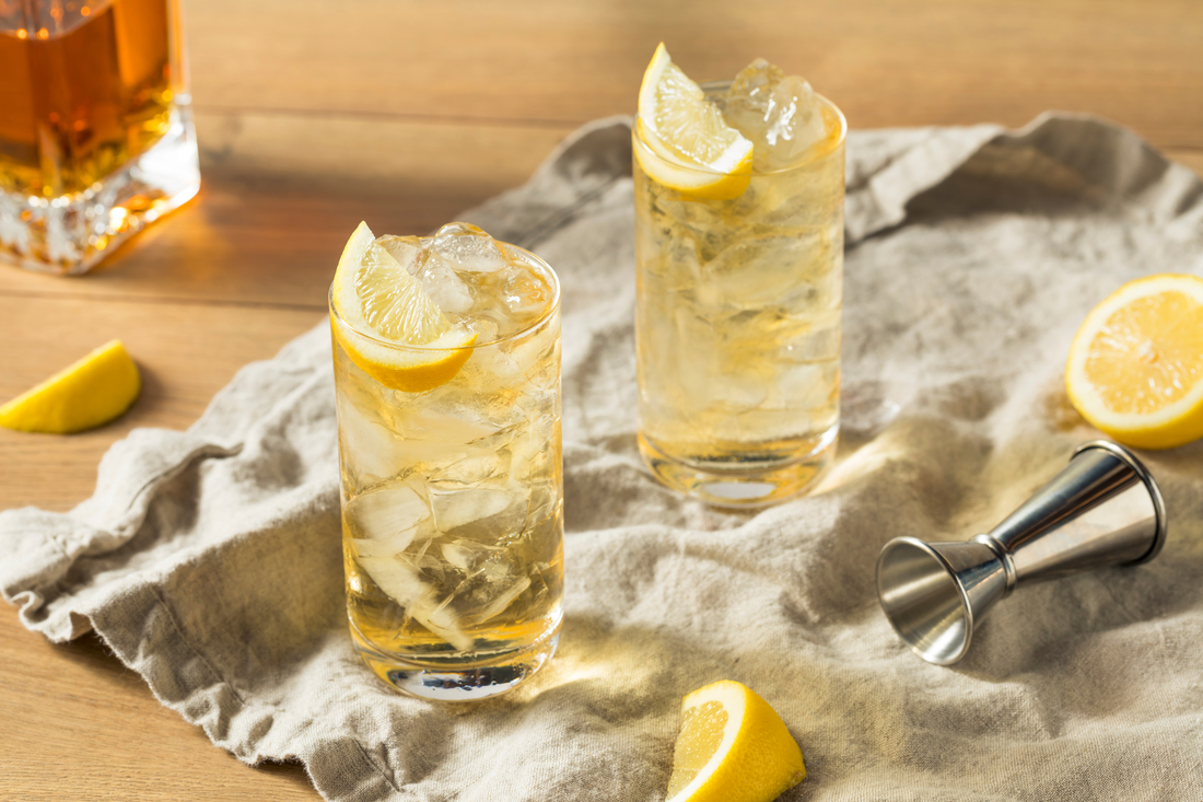 Two highball glasses filled with iced tea, ice cubes, and lemon wedges on a cloth napkin, with a jigger, lemon slices, and a bottle in the background.