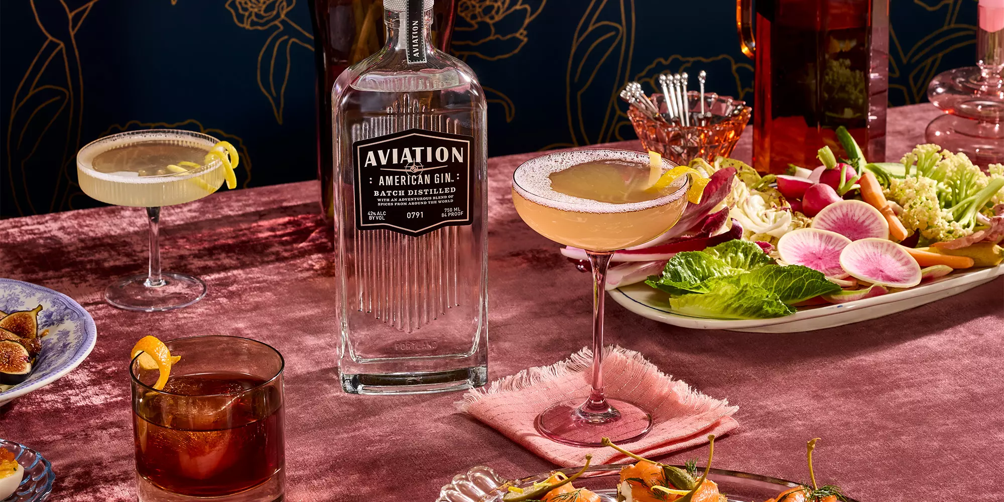 A bottle of Aviation American Gin is surrounded by cocktails, garnished drinks, and a platter of fresh vegetables on a pink velvet tablecloth.