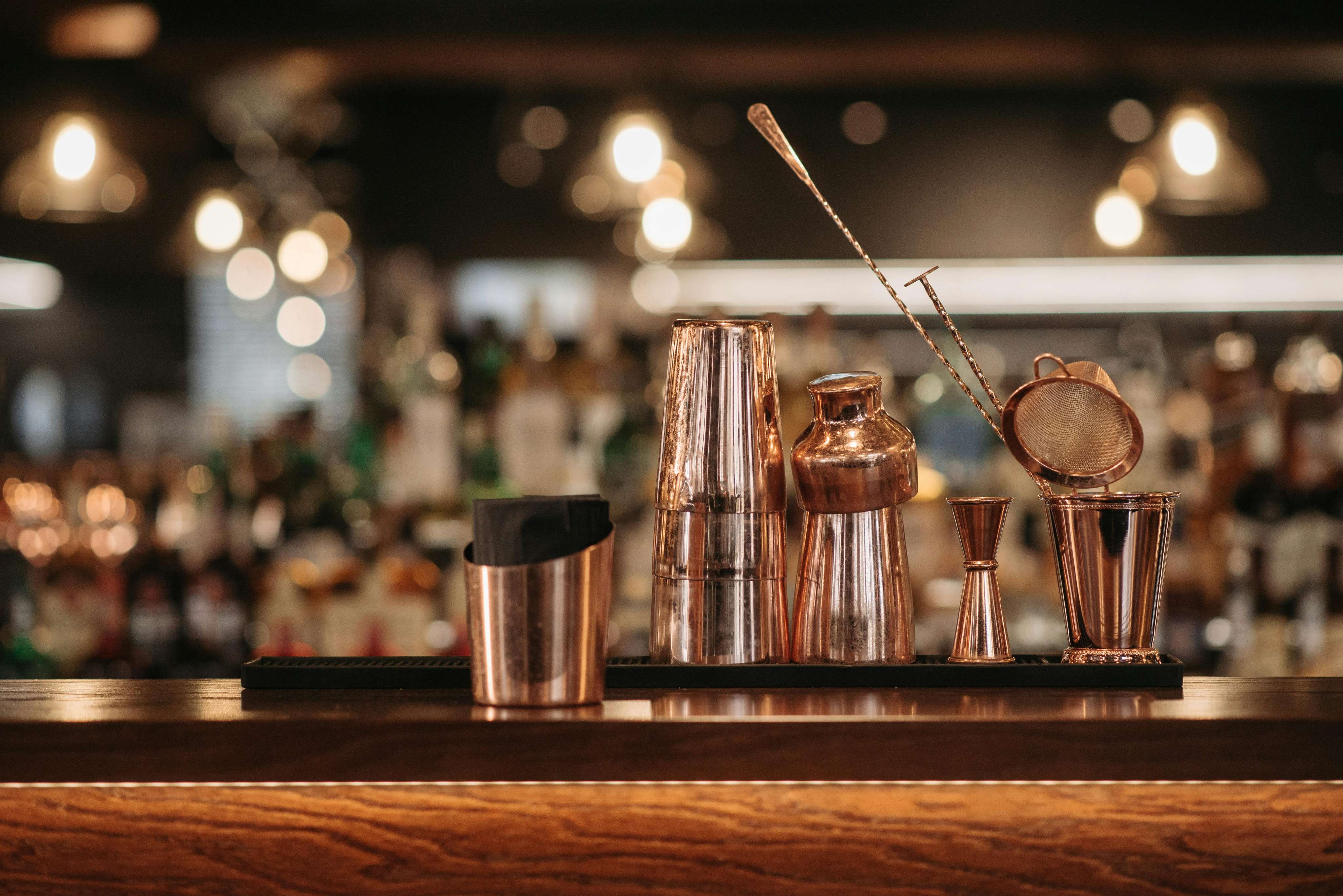 A set of copper bar tools, including a shaker, jigger, strainer, and mixing spoon, arranged on a wooden bar counter with blurred bottles in the background.