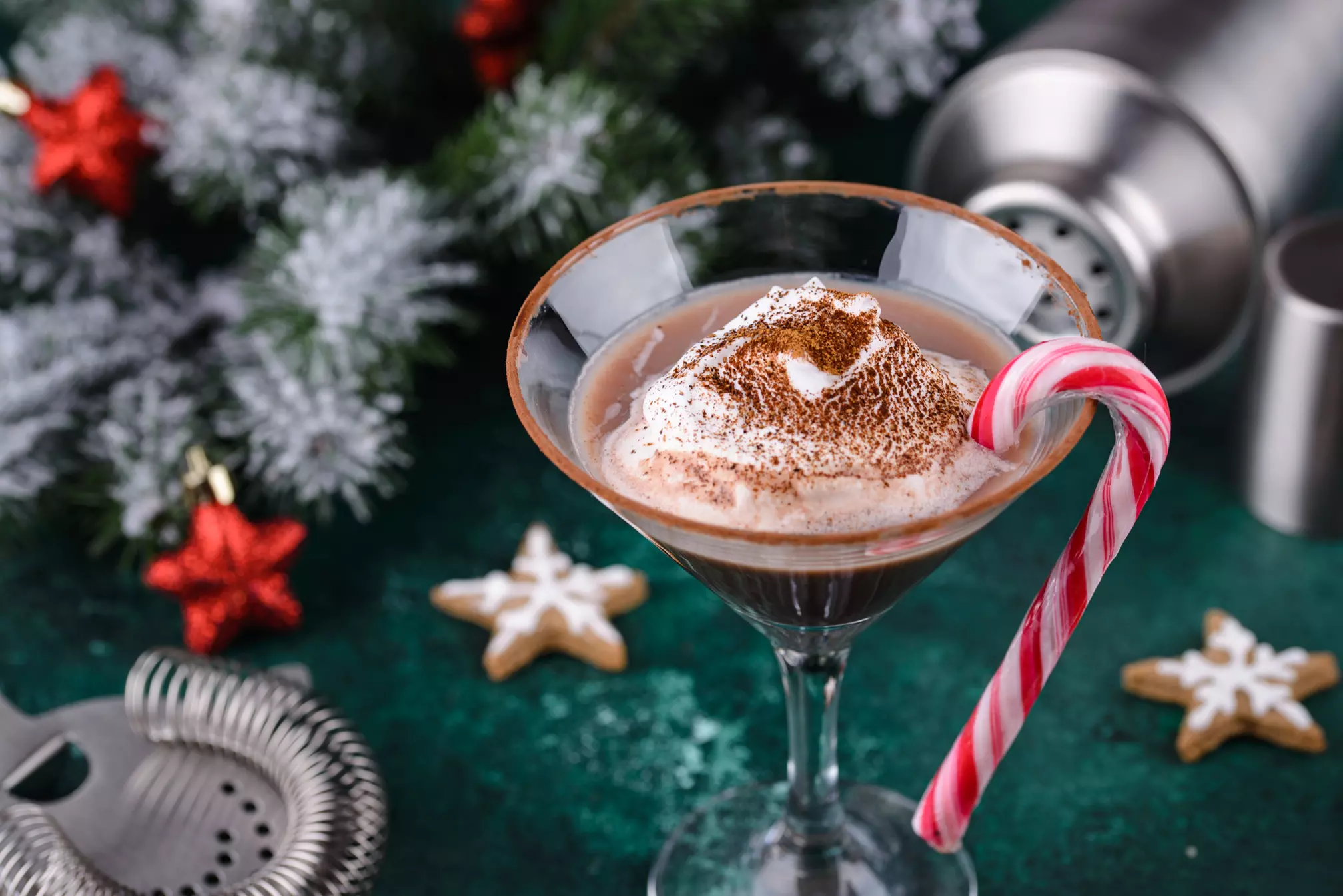 A chocolate cocktail in a martini glass topped with whipped cream and cocoa, garnished with a candy cane. Christmas decorations and gingerbread cookies are in the background.