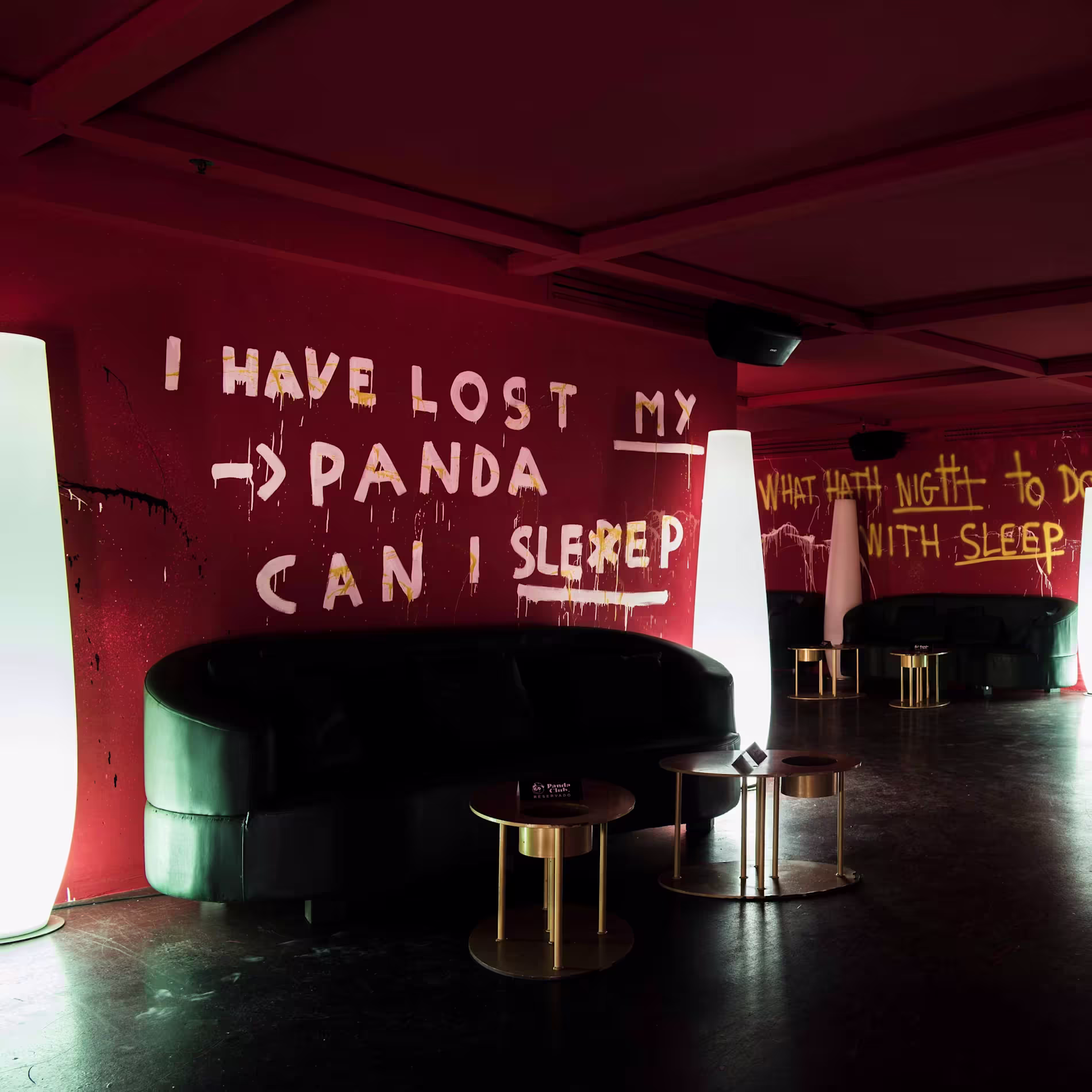 Panda Club: Un espacio intrigante con paredes rojas cubiertas de grafitis artísticos y frases misteriosas. La iluminación tenue y los sofás negros curvos crean un ambiente íntimo y moderno.