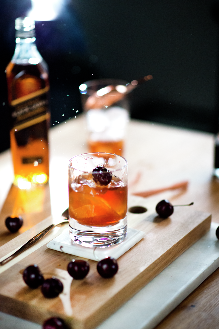 La imagen muestra un vaso relleno con el cóctel Manhattan, elementos de coctelería y, de fondo, se puede visualizar la botella de whisky Johnnie Walker Black Label.