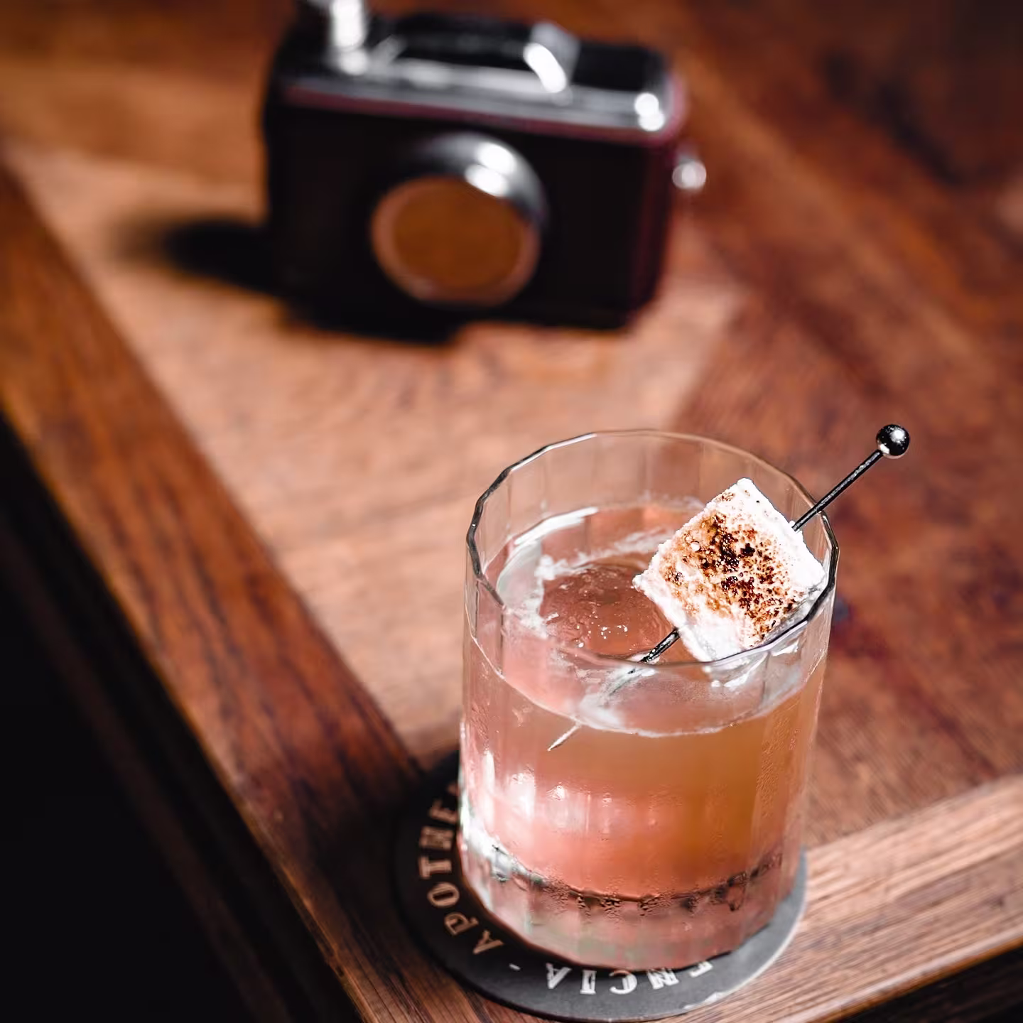Cóctel de autor en Apotheke con hielo esférico y nube tostada, servido sobre una base de madera con una cámara vintage al fondo.