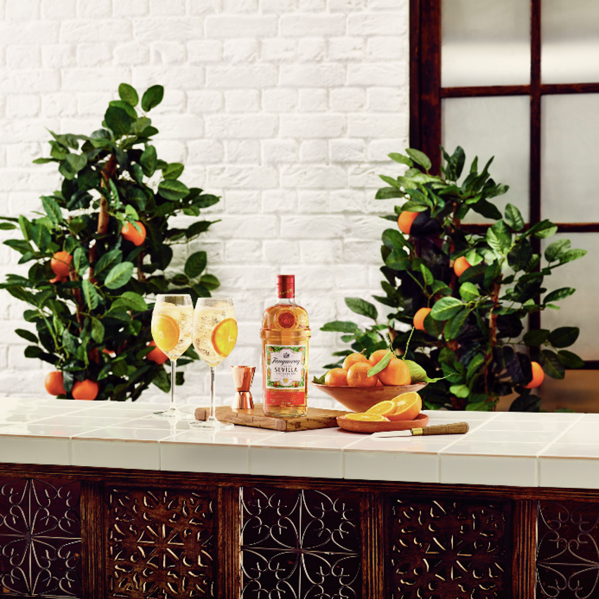La imagen muestra una botella de gin Tanqueray Flor de Sevilla, dos copas con cócteles preparados con dicha bebida y elementos de coctelería sobre una mesada. De fondo, se observan dos árboles de naranja.