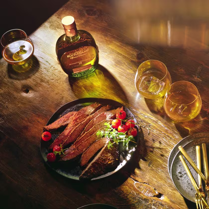 La imagen muestra la botella de Buchanan's Special Reserve junto a un plato de comida con carne y tres copas llenas de este whisky.