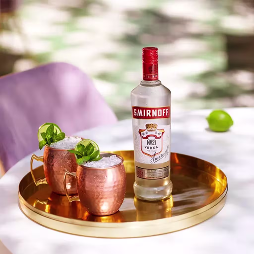 Bottle of smirnoff no 21 with two copper cups of smirnoff vodka on ice, garnished with mint leaves on a copper tray