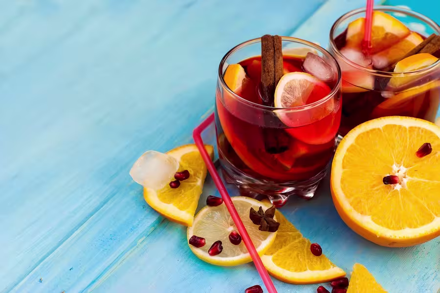 Two glasses of iced fruit punch with orange slices, cinnamon sticks, and star anise, surrounded by orange slices, ice cubes, and pomegranate seeds on a blue wooden surface.