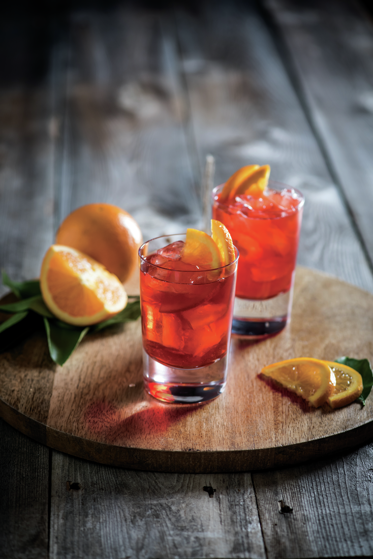 La imagen muestra dos vasos rellenos con el cóctel Negroni y naranjas alrededor.