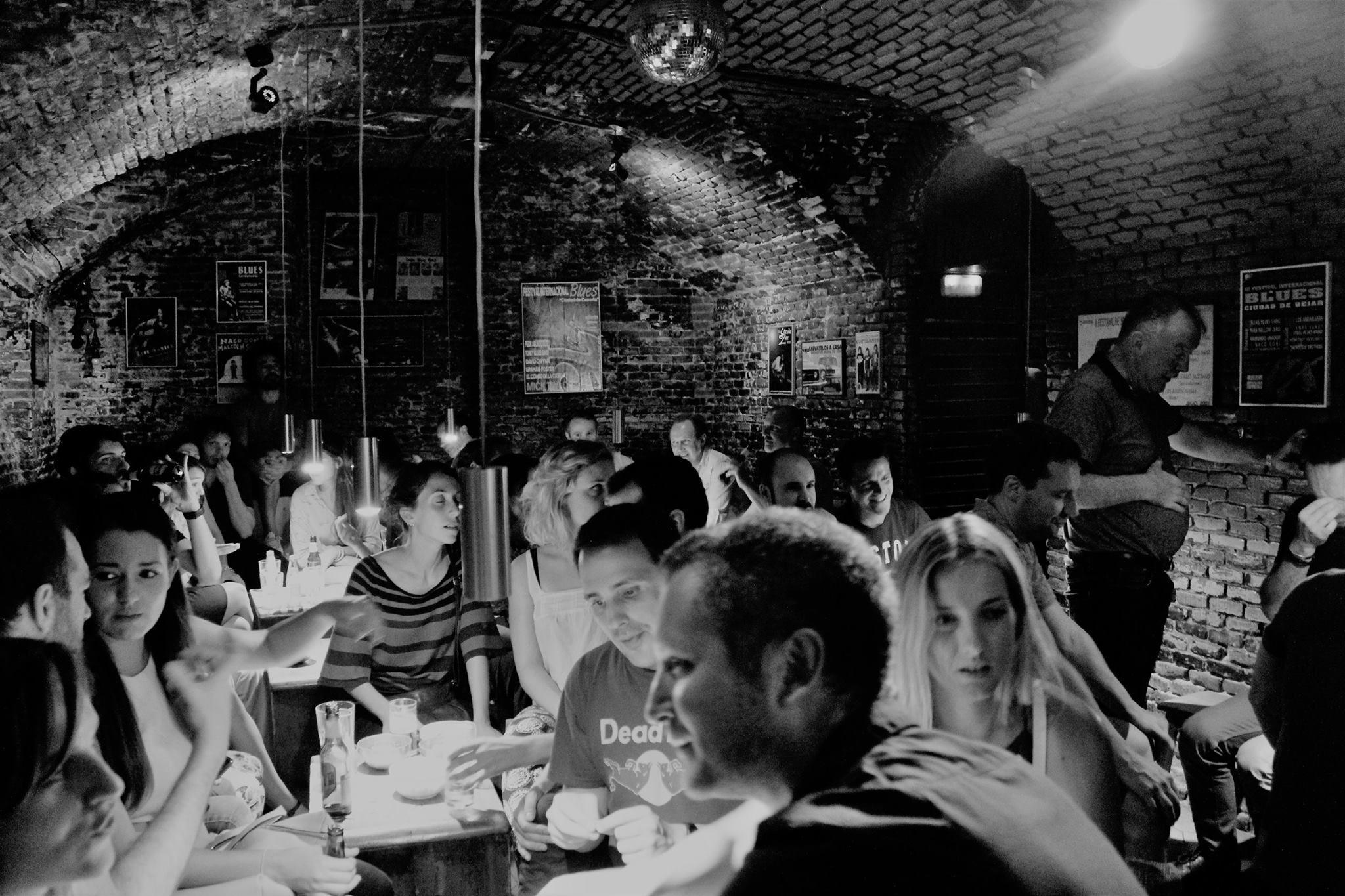 La foto muestra un espacio interior de La Coquette, con paredes de ladrillo y techos arqueados, creando una atmósfera acogedora. Hay varias personas sentadas en mesas, algunas con bebidas frente a ellas.