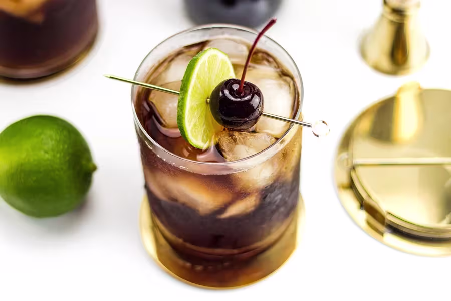 A glass of dark iced cocktail garnished with a lime slice and cherry on a cocktail pick, placed on a gold coaster with a lime and bar tools nearby.