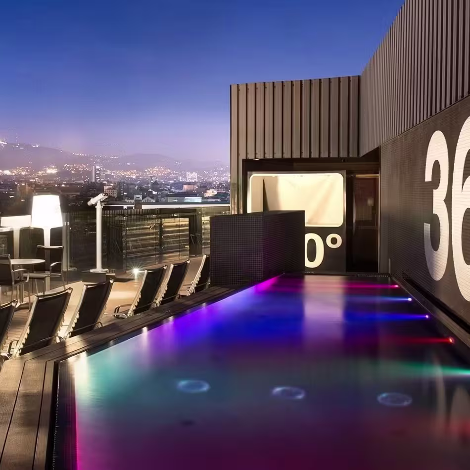  Terraza del Barcelo Raval, con vista a un paisaje urbano en el fondo. El área está equipada con muebles modernos, incluyendo mesas y sillas, y cuenta con una piscina iluminada con colores cambiantes. En la pared se muestra prominentemente el logo “360°”.