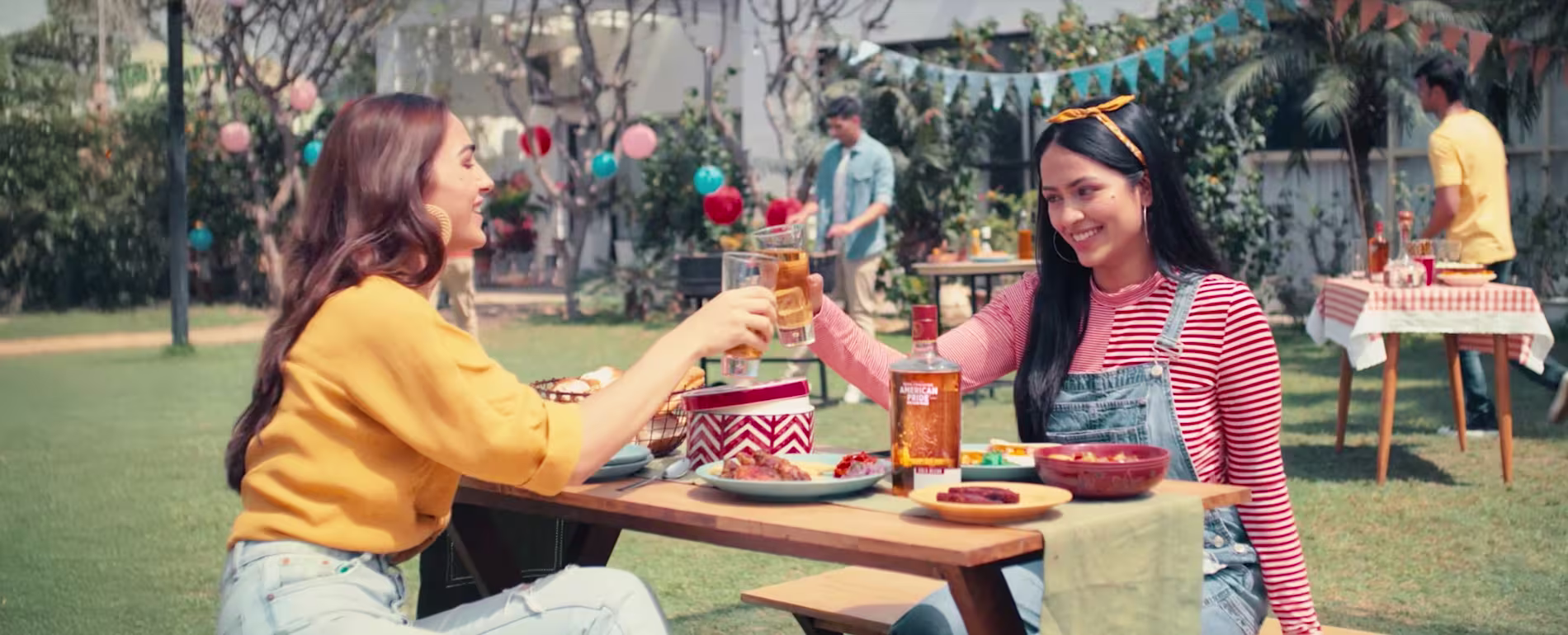 Two women sit at an outdoor table, smiling and clinking glasses, with food and a bottle of whiskey visible. Other people and picnic tables are in the background.