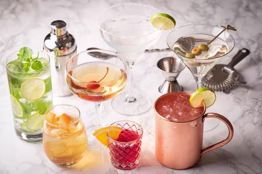 A variety of cocktails in different glasses, including a copper mug, garnished with lime, mint, olives, orange, and cherries, arranged on a marble surface with bar tools.