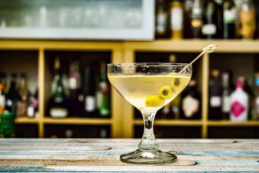 A martini garnished with green olives on a toothpick sits on a wooden surface, with shelves of bottles in the blurred background.