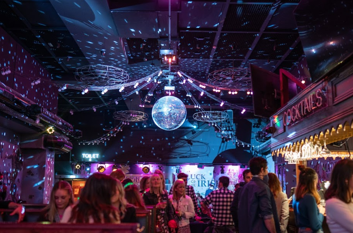 The Old Irish Pub destaca por su ambiente vibrante con una bola de discoteca iluminada y luces de neón, creando la atmósfera perfecta para la fiesta.