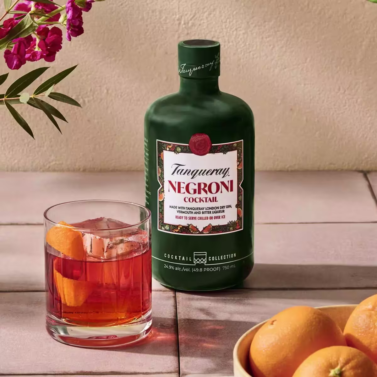 A bottle of Tanqueray Negroni Cocktail stands next to a glass of Negroni with orange garnish and a bowl of oranges on a tiled surface.