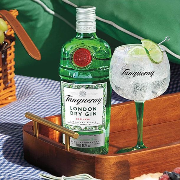 Egy üveg Tanquery London Dry Gin ül egy fatálcán egy márkás Tanqueray pohár mellett, amely gint és tonikot tartalmaz.